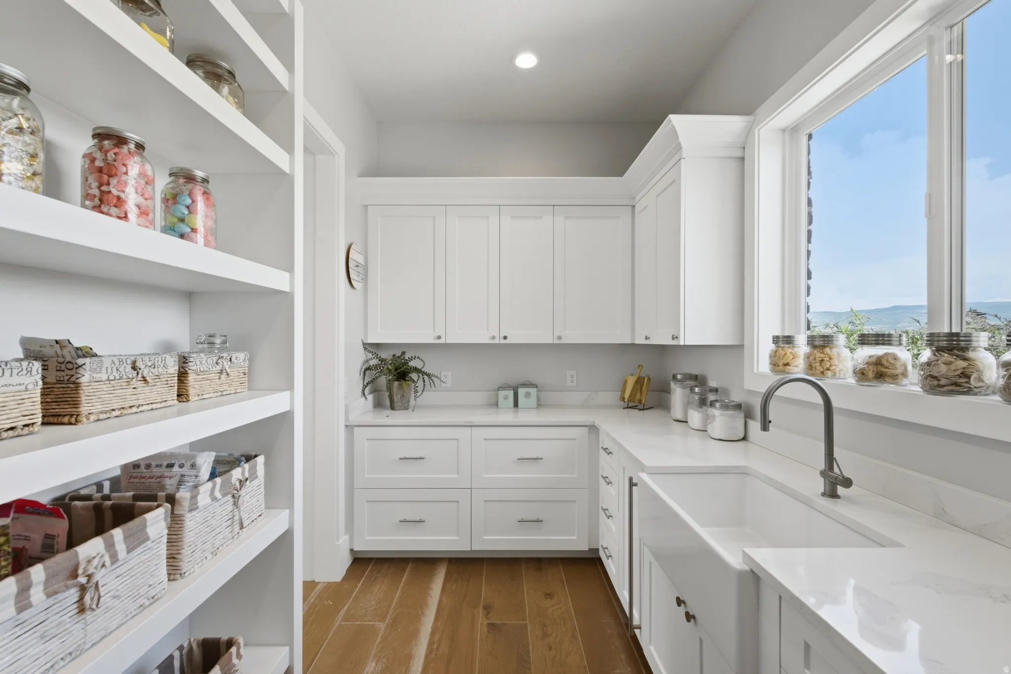 Well-appointed pantry featuring extensive built-in shelving, white shaker cabinetry, an integrated farmhouse sink with a gooseneck faucet, light-toned countertops, and wood-finish flooring