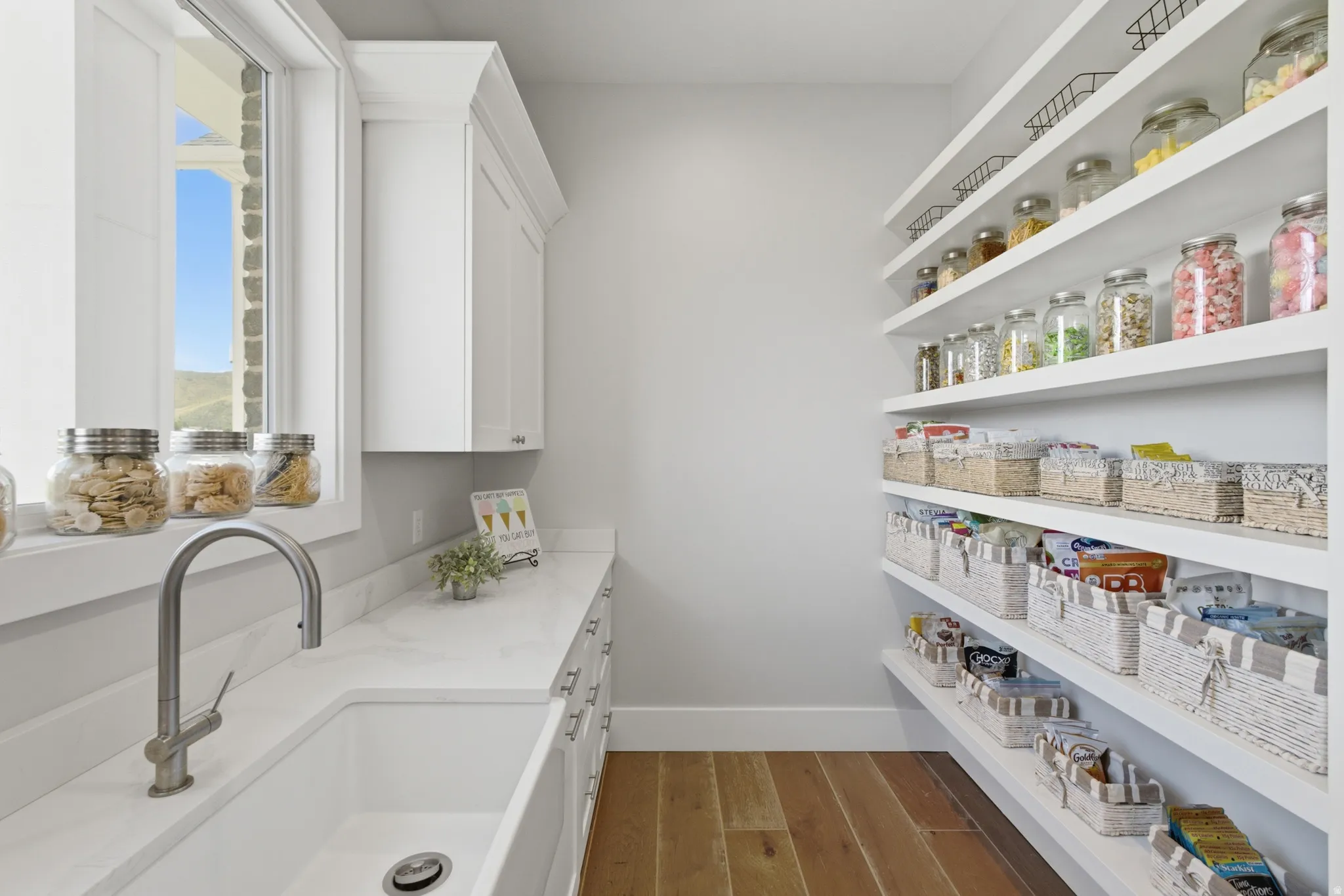 Bright pantry featuring extensive open shelving, wood-finish flooring, white cabinetry, a large sink, and a window