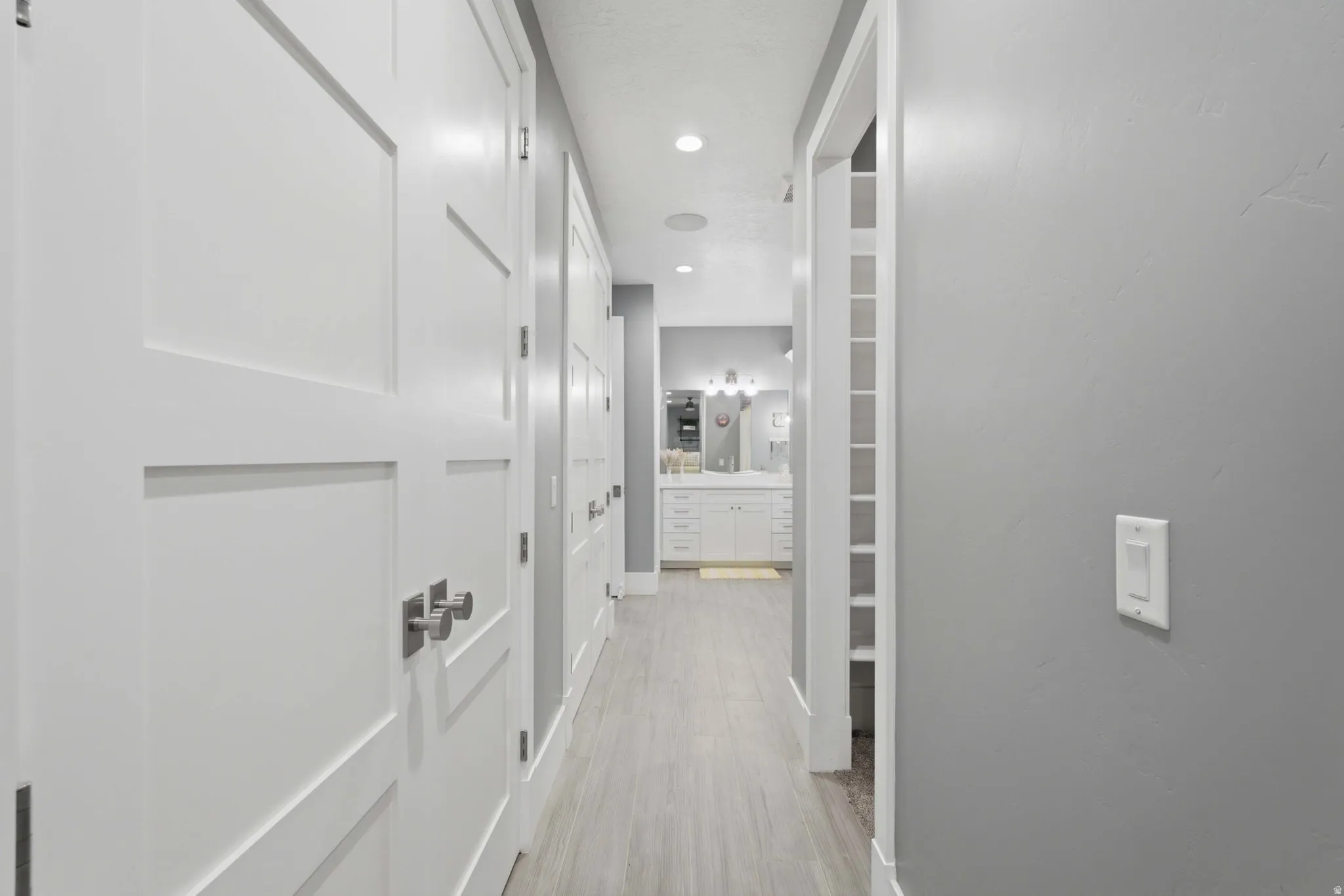 Hallway featuring light wood-finish flooring, white paneled doors with brushed nickel hardware, light grey painted walls, and recessed lighting