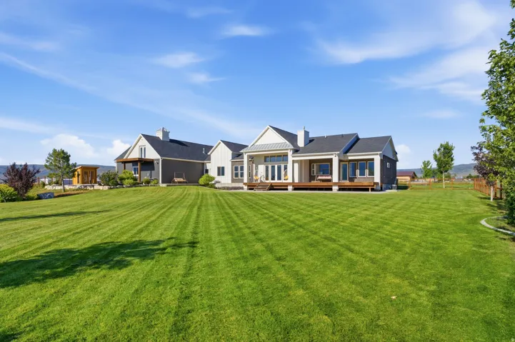 Expansive green lawn with a contemporary multi-gabled home featuring a covered porch, outdoor decking, multiple windows, and a partially fenced perimeter