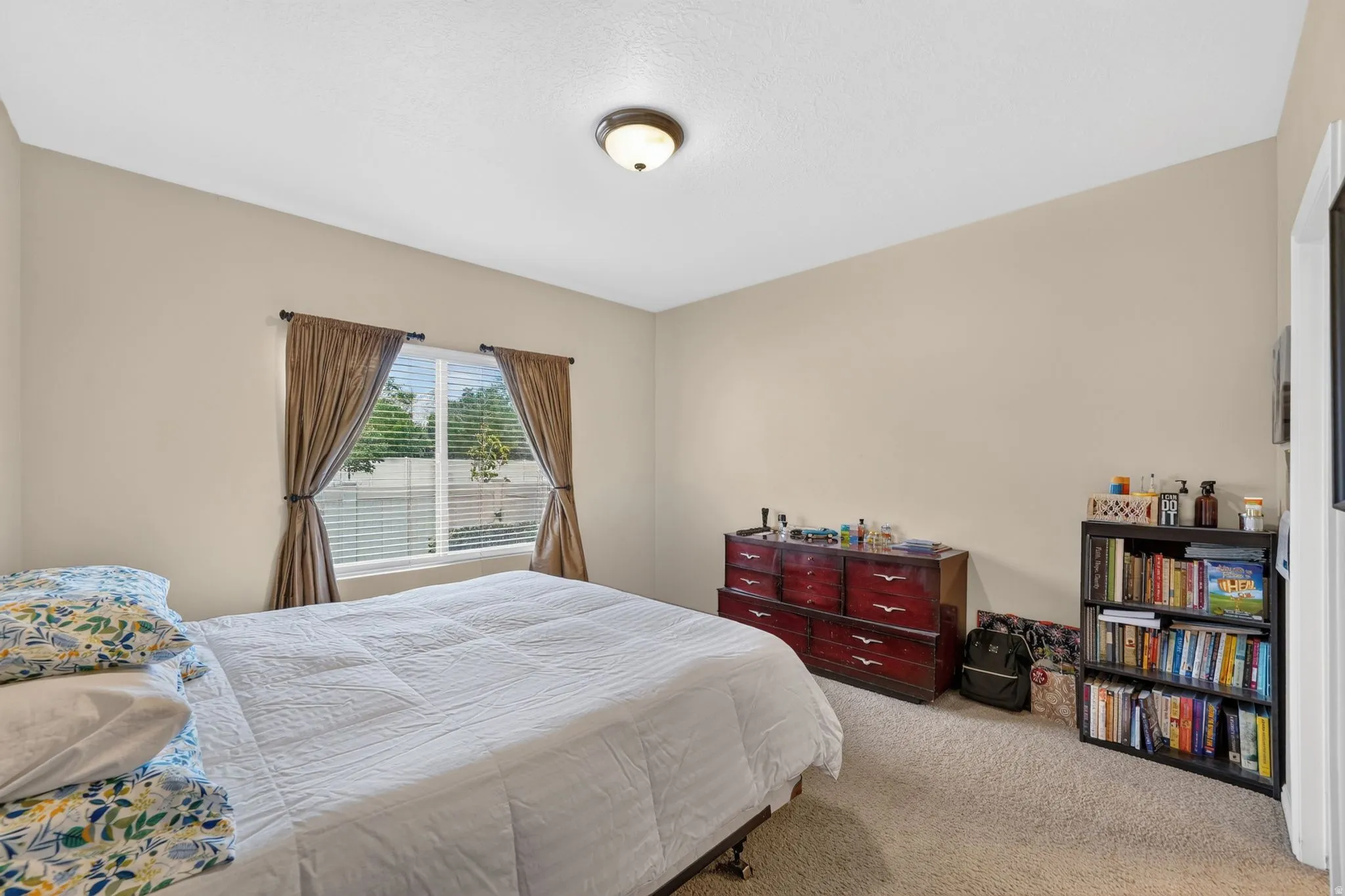 Bedroom featuring a window with blinds and drapes, neutral-toned walls, textured carpeting, and a flush-mount ceiling light fixture