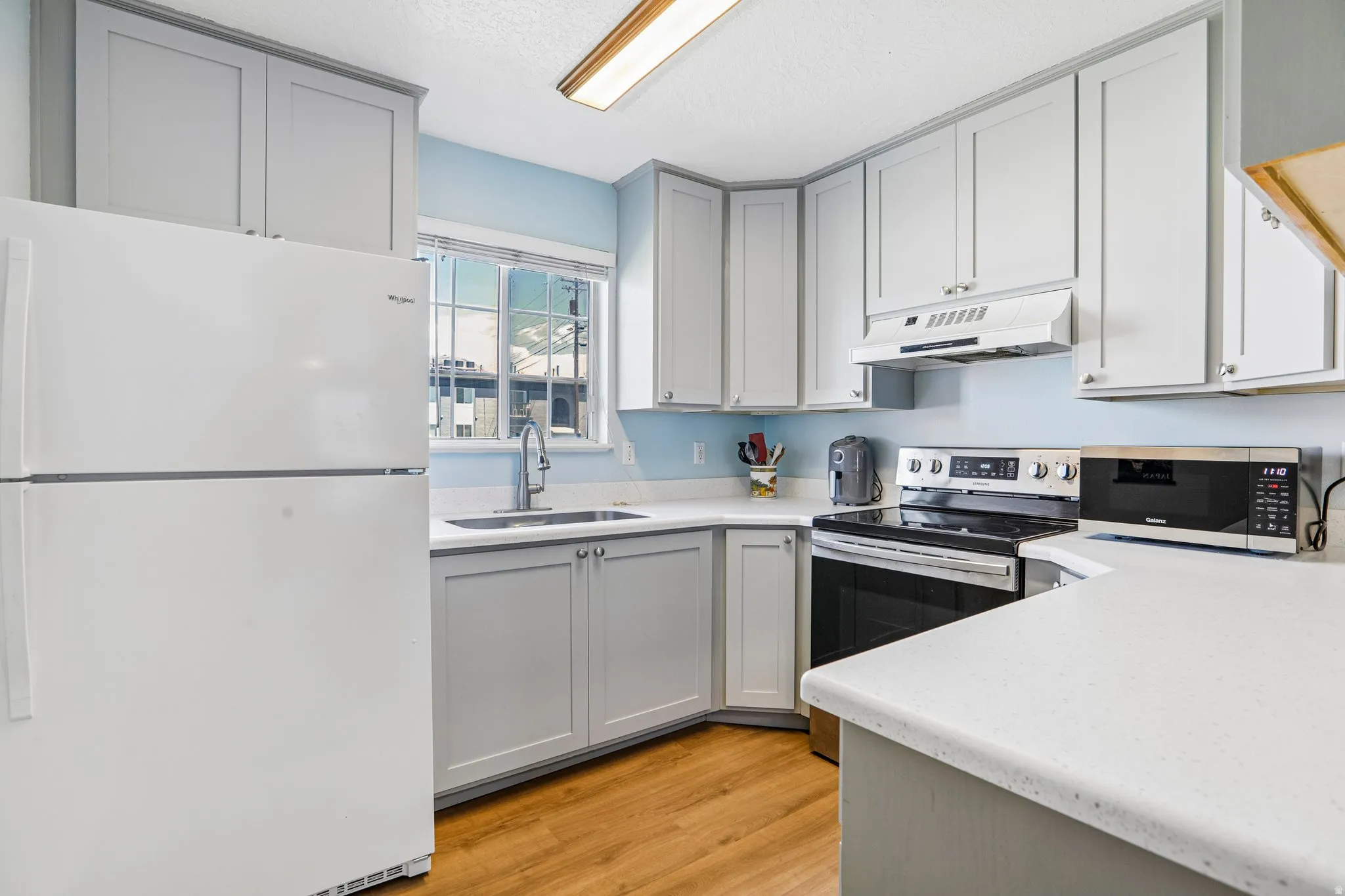 Kitchen featuring light gray cabinetry, white countertops, stainless steel appliances, wood-finish flooring, and a window above the sink