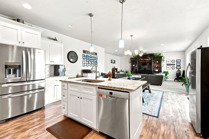 Kitchen and living area featuring wood-finish flooring, white cabinetry, stainless steel appliances, a center island with a sink and light-toned countertop, and recessed lighting