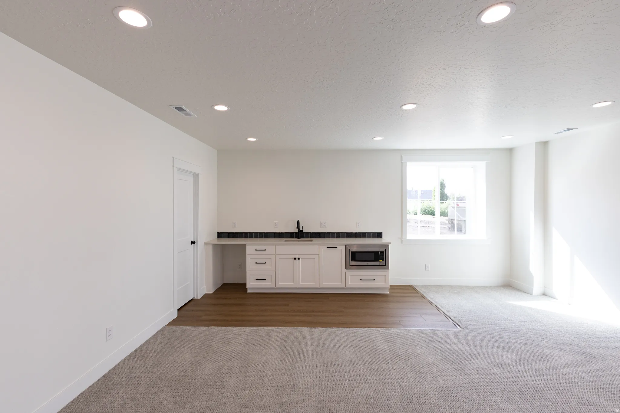 Spacious room featuring recessed lighting, a built-in wet bar with a stainless steel microwave, white cabinetry, dark hardware, and a window