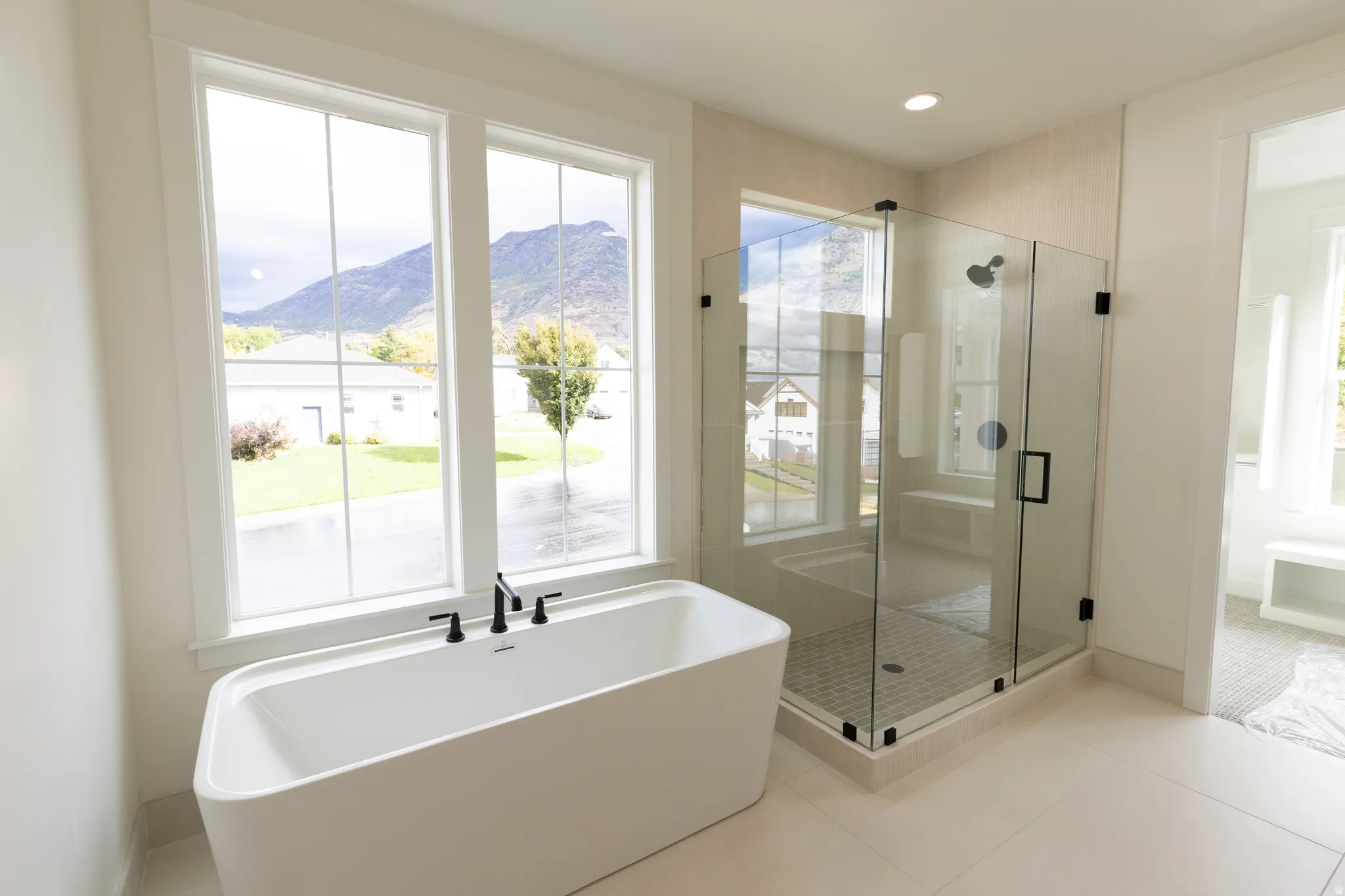 Freestanding tub with a matte black faucet, glass-enclosed shower with black hardware, large format floor tiles, and abundant natural light from multiple windows