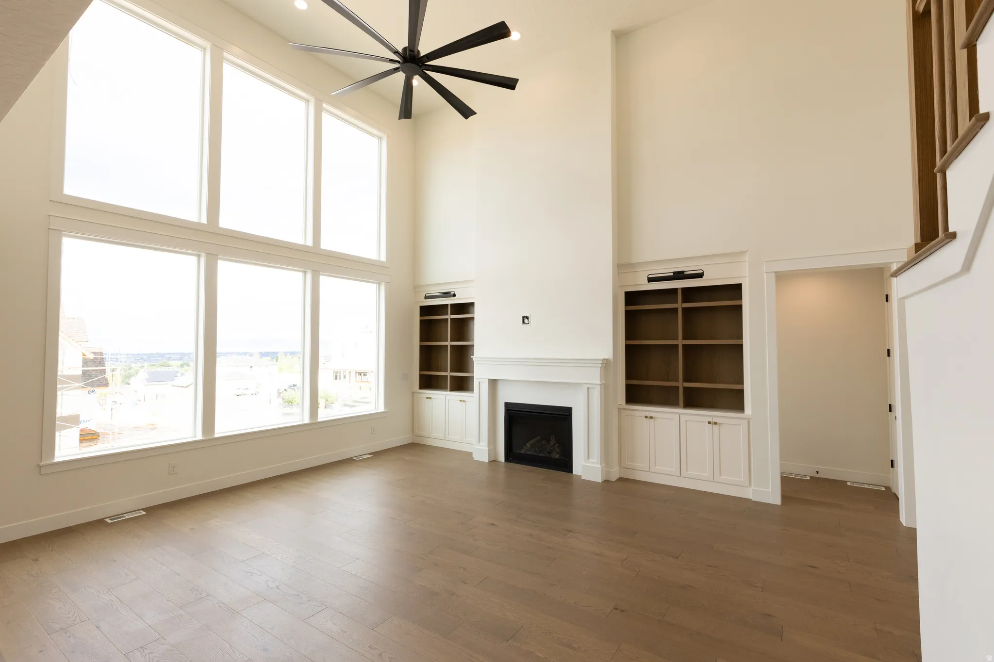 Expansive living space featuring a soaring ceiling, large windows, wood-finish flooring, a contemporary ceiling fan, and built-in shelving with cabinetry