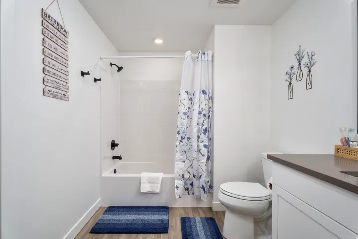 Bathroom featuring a white bathtub with subway tile surround, matte black shower fixtures, wood-finish flooring, a white vanity with a dark countertop, and a white toilet