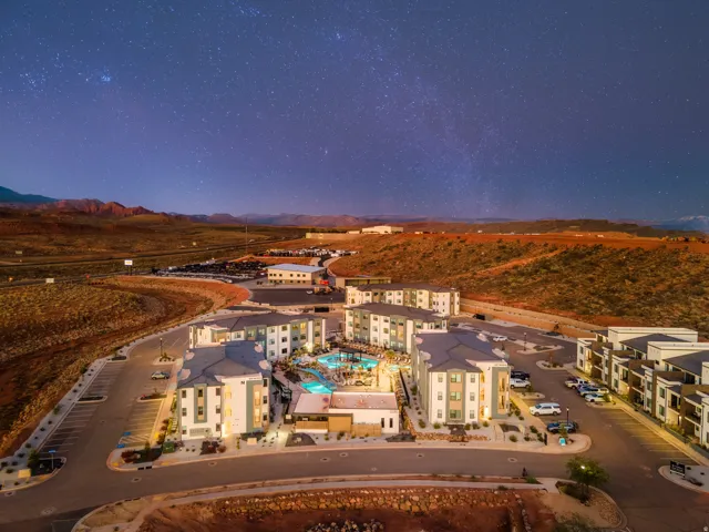 Modern multi-story buildings featuring a central swimming pool with integrated lighting, surrounding paved patios, and dedicated parking areas