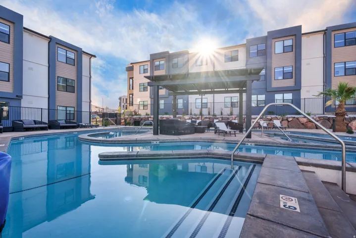 Expansive community pool area featuring an integrated spa, surrounding lounge seating, a shaded pergola, and a rock-landscaped perimeter