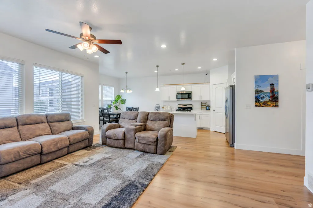 Bright living space featuring wood-finish flooring, a ceiling fan with integrated lighting, and multiple windows with horizontal blinds