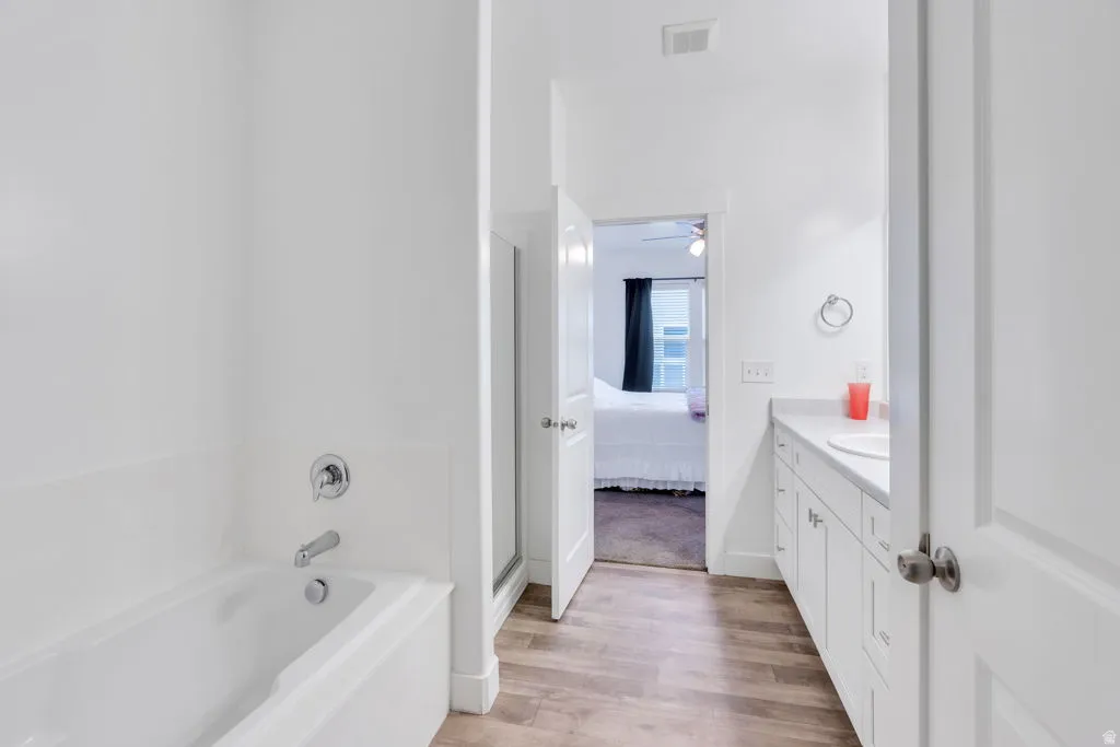 Modern bathroom featuring a white soaking tub with chrome fixtures, a separate glass-enclosed shower, a long vanity with dual white shaker cabinets, a single integrated sink, and wood-finish flooring