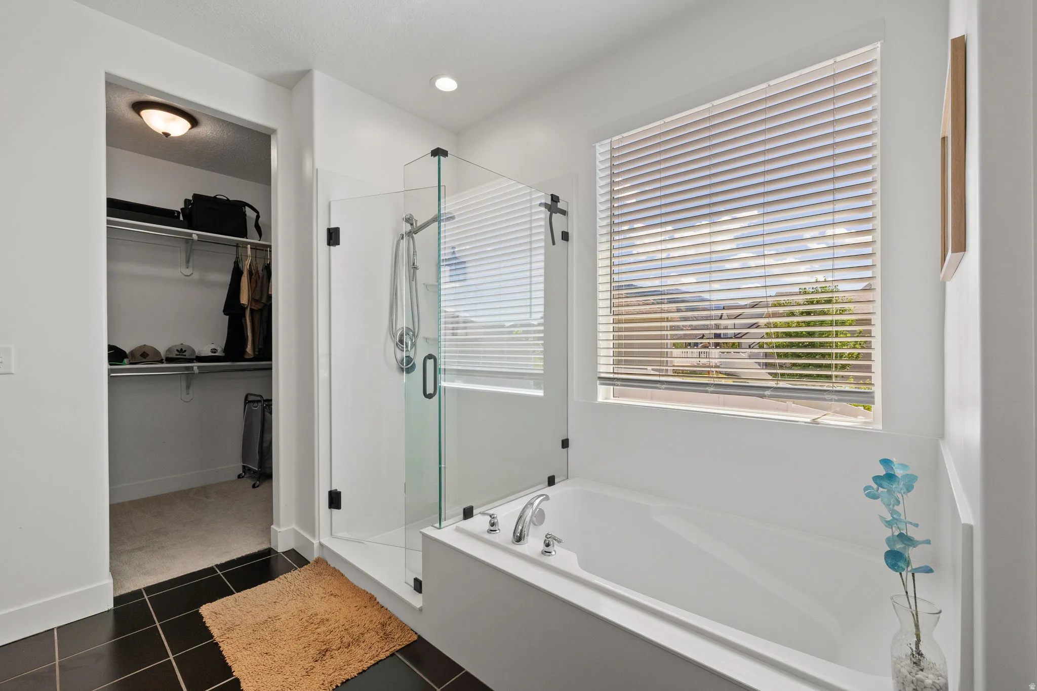 Bathroom featuring a built-in soaking tub, glass-enclosed shower, dark tile flooring, and an adjacent walk-in closet with shelving and hanging rods