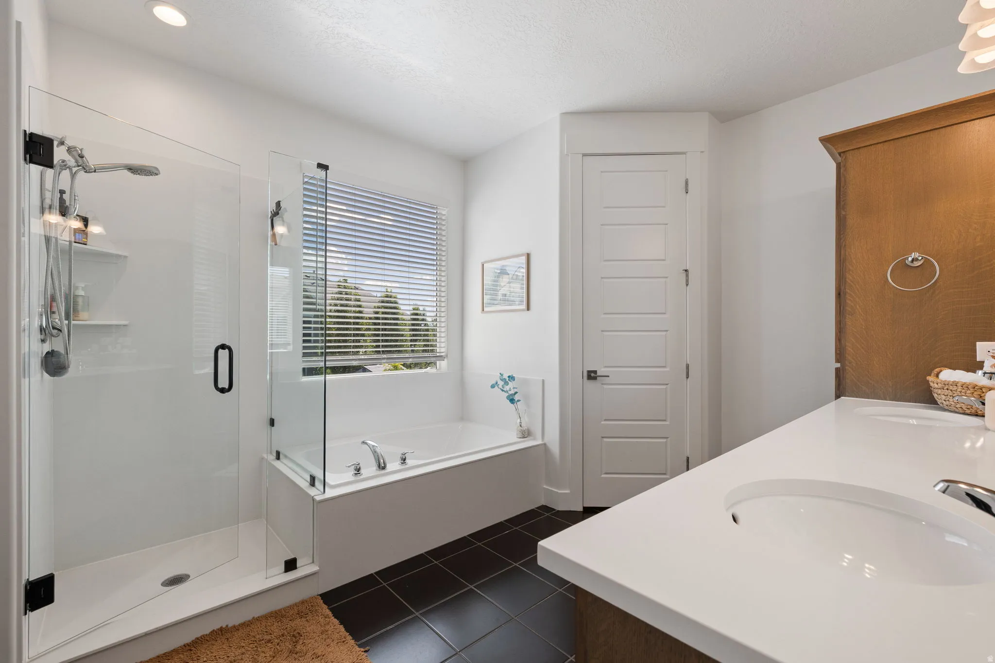 Bathroom featuring a frameless glass shower, a soaking tub with a window, dark tile flooring, a dual-sink vanity with white countertops, and a paneled interior door