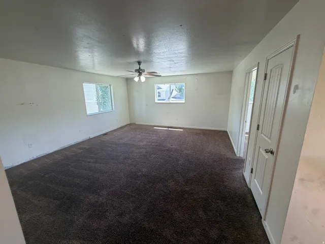 Spacious room featuring neutral-toned walls, dark patterned carpeting, and two windows providing natural illumination