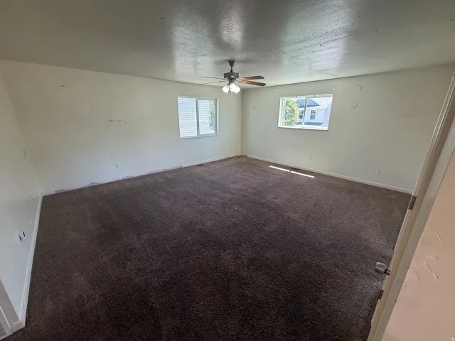 Expansive room with neutral-toned walls, brown carpeting, and a ceiling fan with integrated lighting