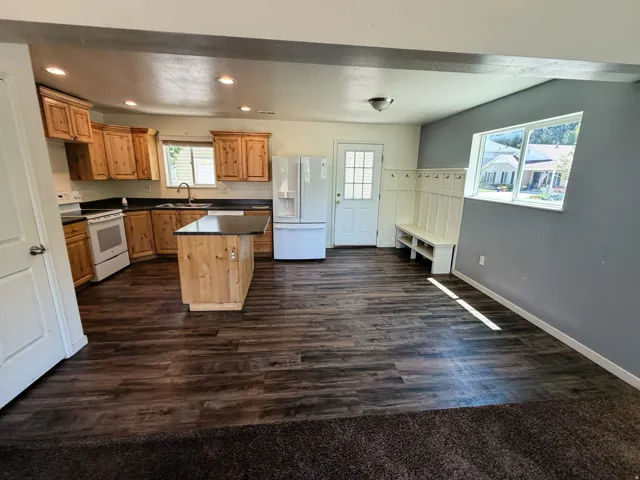 Open-concept kitchen and entryway featuring wood-finish flooring, light wood cabinetry, white appliances, a kitchen island with seating potential, and a built-in hall tree with bench storage