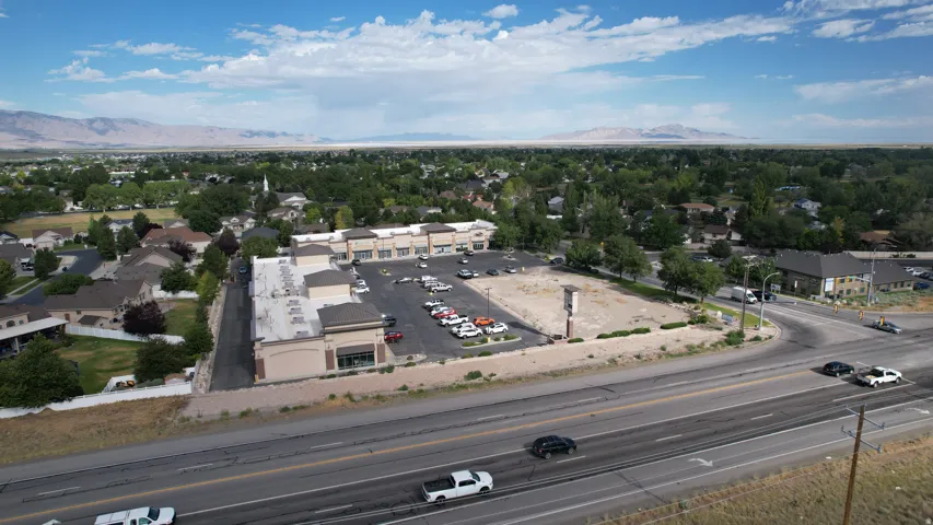 Commercial retail center featuring a large asphalt parking lot, multiple storefronts with neutral-toned facades, and prominent road frontage