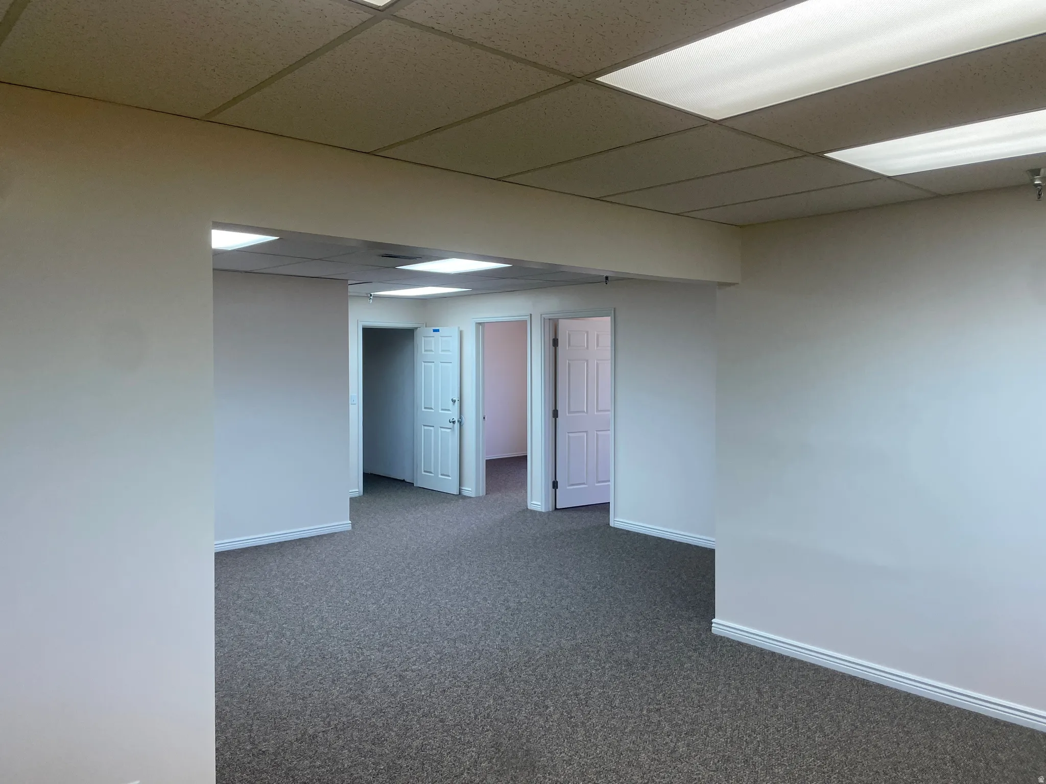 Open commercial space featuring neutral-toned walls, gray patterned carpeting, and a drop ceiling with integrated fluorescent lighting