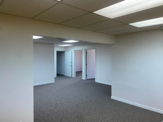 Open commercial space featuring neutral-toned walls, gray patterned carpeting, and a drop ceiling with integrated fluorescent lighting