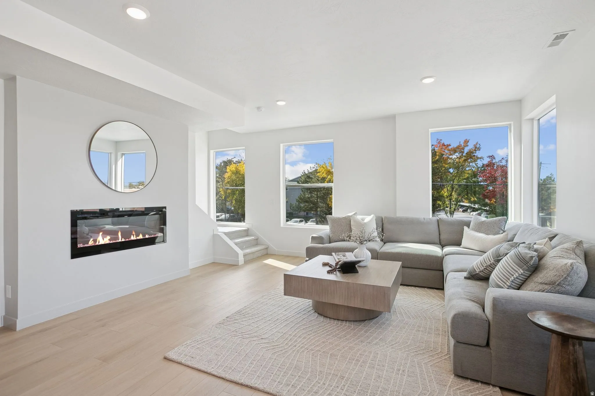 Open-concept living space with light wood-finish flooring, a contemporary wall-mounted fireplace, recessed lighting, and multiple large windows