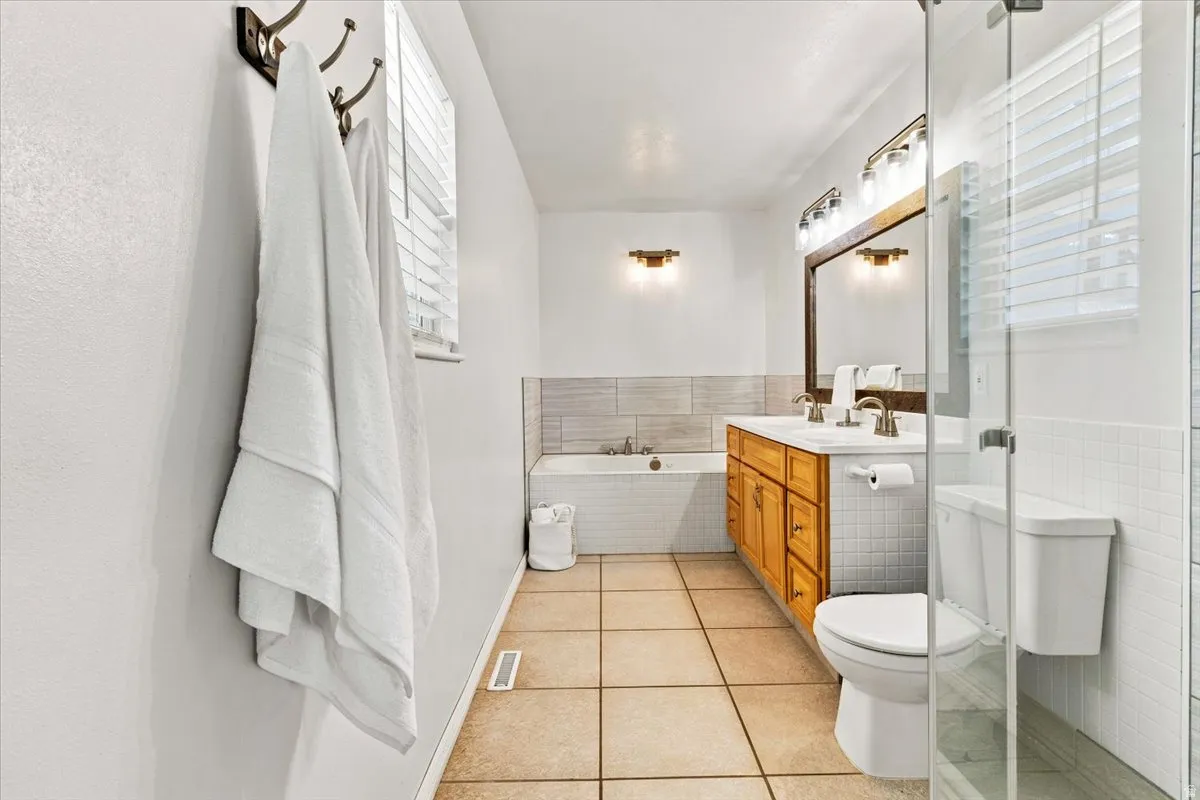 Bathroom featuring a dual vanity with wood cabinetry, a built-in tub with tile surround, a separate shower with a glass enclosure, and light-toned tile flooring