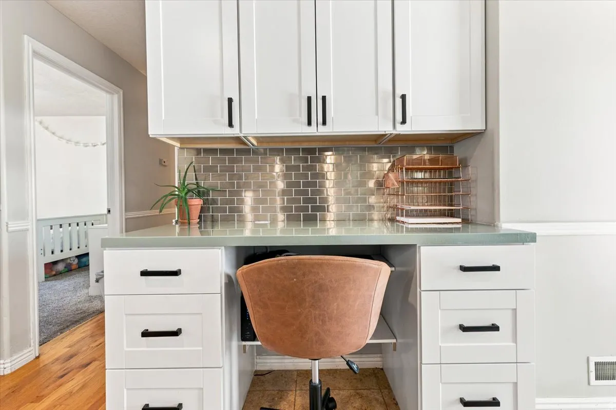 Built-in desk featuring a light-toned countertop, white cabinetry with black hardware, and a metallic subway tile backsplash