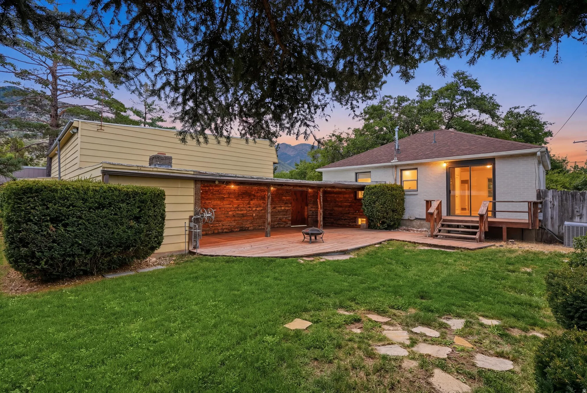 Expansive backyard featuring a manicured lawn, a large wood deck with steps, a covered patio with exposed wood beams and string lighting, and a sliding glass door leading into the residence