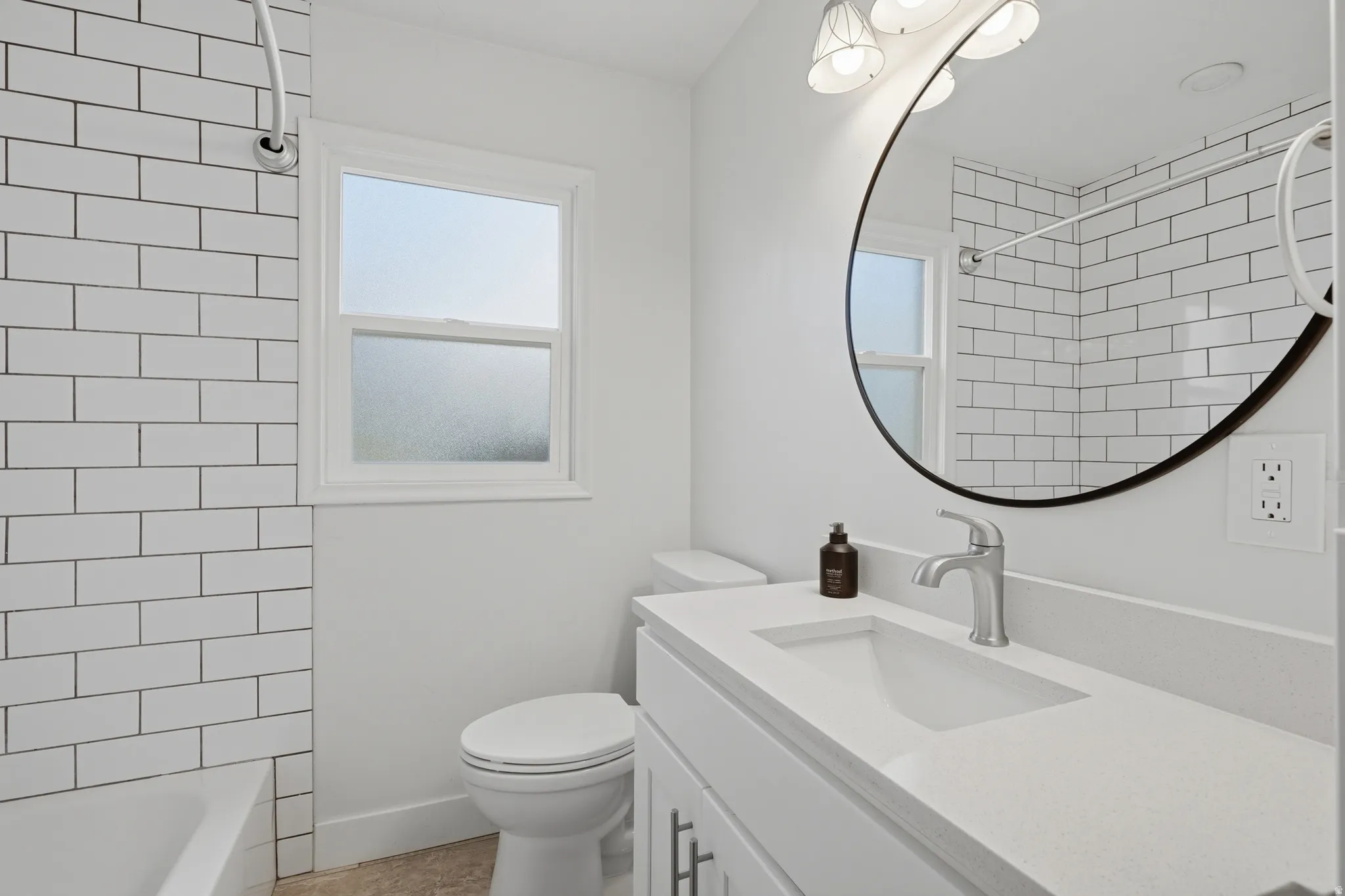 Bathroom featuring subway tile shower surround, white vanity with integrated sink, circular mirror, and window with privacy glass