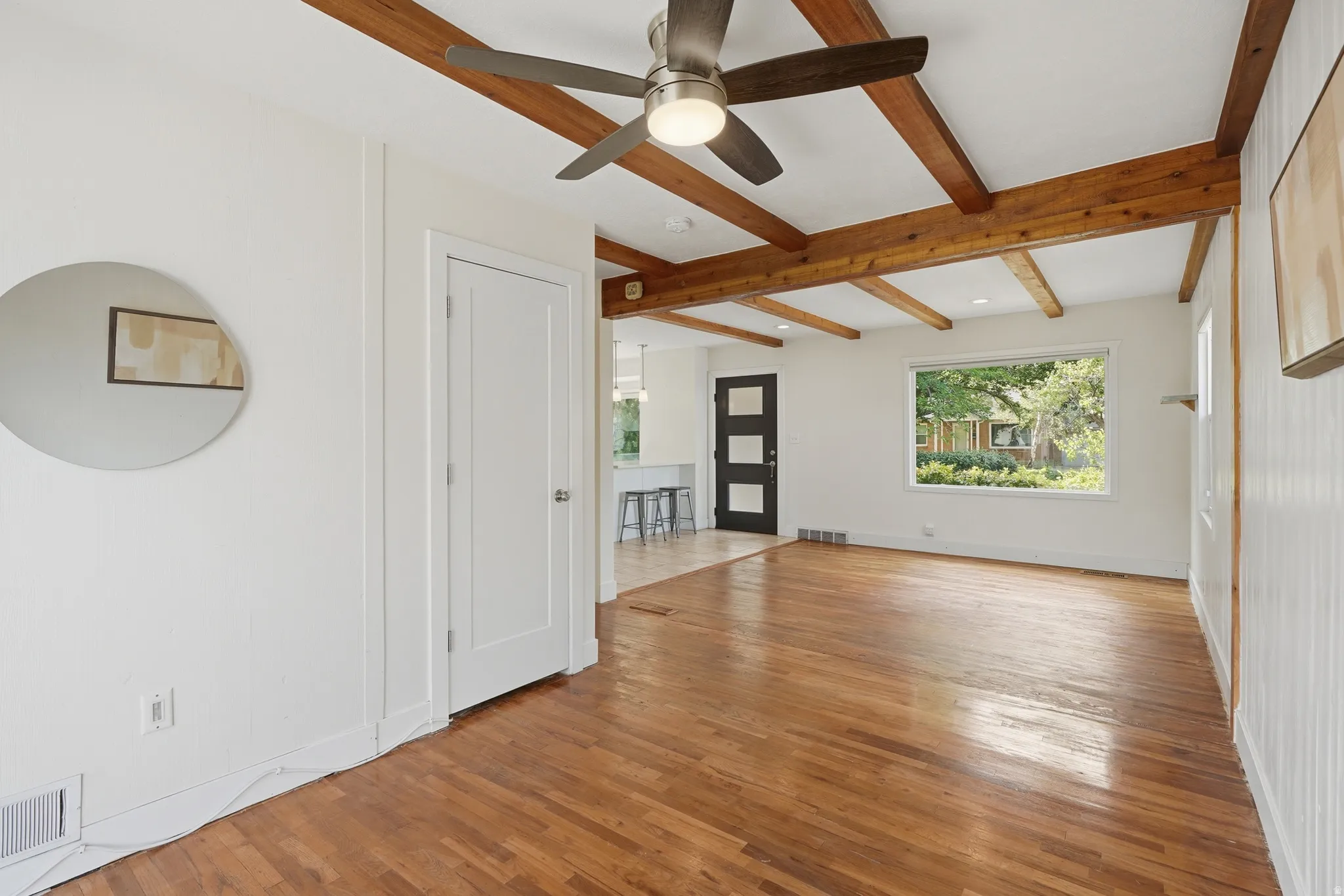 Exposed wood ceiling beams enhance this expansive room featuring wood-finish flooring, a contemporary ceiling fan, and a large window
