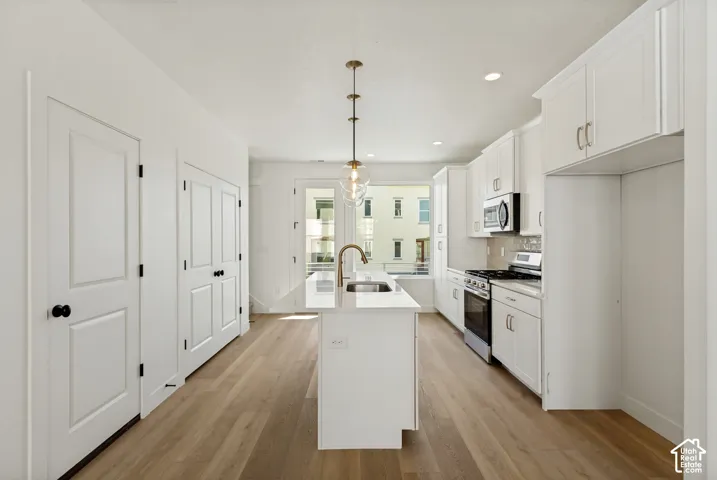 Kitchen featuring white shaker cabinetry, stainless steel appliances, wood-finish flooring, a center island with sink, and a pendant light fixture
