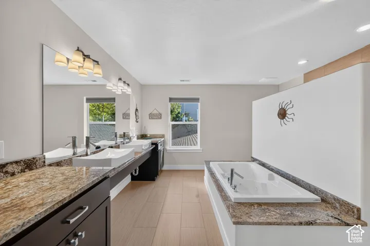 Expansive bathroom featuring a built-in soaking tub with a stone-finish surround, dual vanity with vessel sinks and stone-finish countertops, and wood-finish flooring
