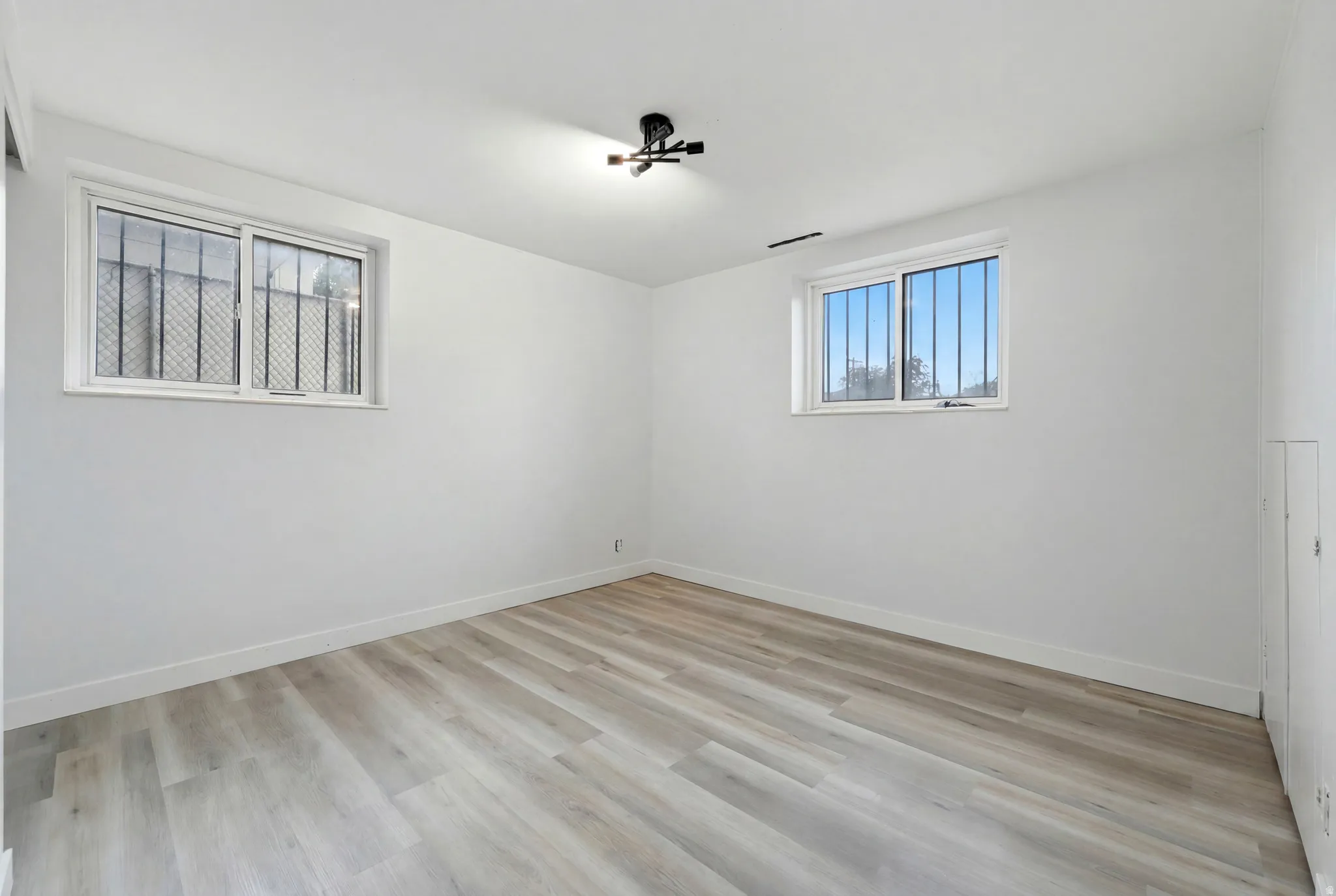 Spacious room featuring light wood-finish flooring, white walls, and two windows with security bars