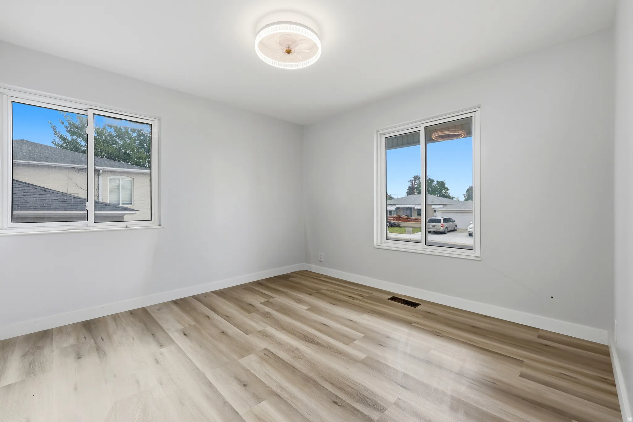Spacious room featuring light gray walls, wood-finish flooring, two large windows, white trim, and a contemporary flush-mount ceiling light