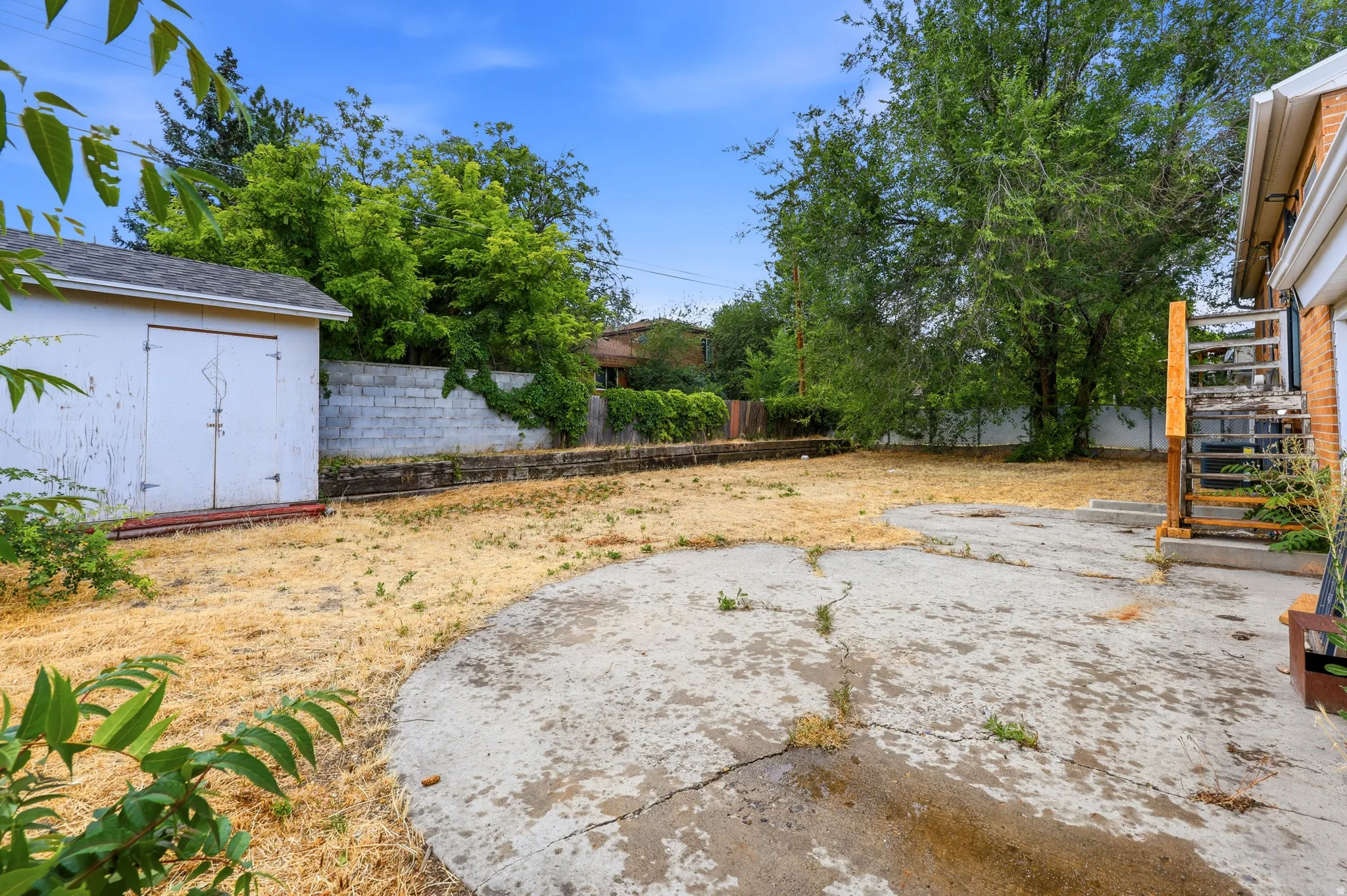 Rear yard featuring a concrete pad, mature trees, a white storage shed, and a concrete block retaining wall