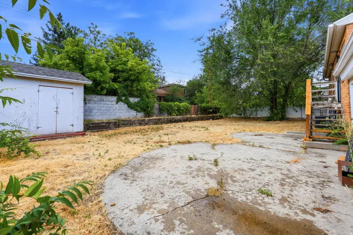 Rear yard featuring a concrete pad, mature trees, a white storage shed, and a concrete block retaining wall