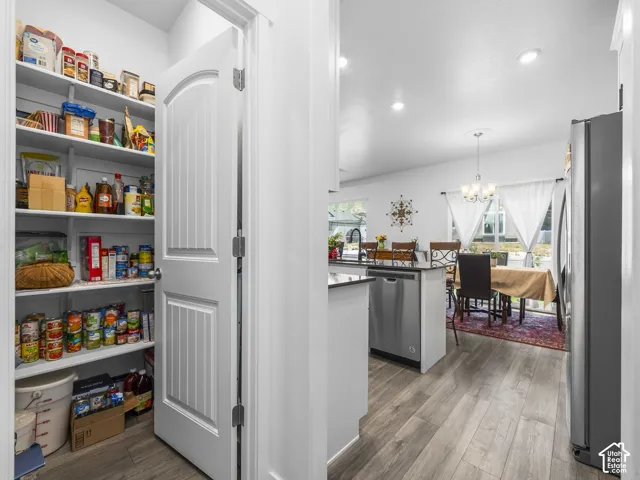 Spacious pantry with multiple shelving rows, complementing the open-concept living area featuring wood-finish flooring, recessed lighting, and a contemporary chandelier
