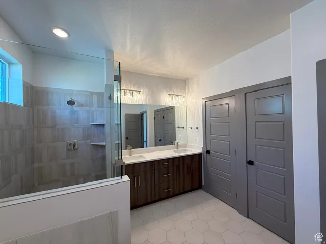 Spacious bathroom featuring a glass-enclosed shower with tile surround and built-in shelving, a dual vanity with a quartz countertop, wood-finish cabinetry, and hexagon tile flooring