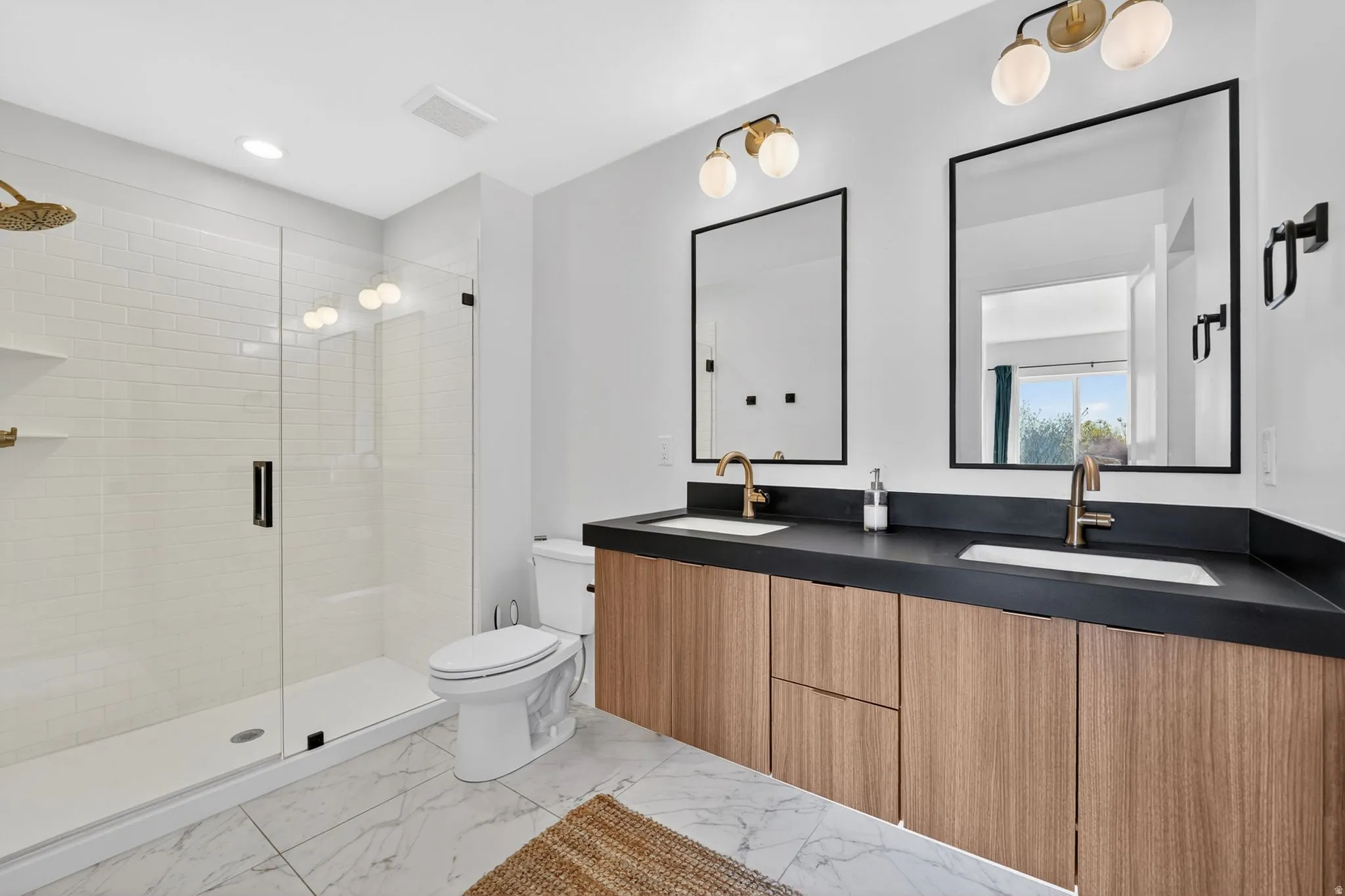 Bathroom featuring a dual vanity with wood-finish cabinetry and dark countertops, two framed mirrors, and a glass-enclosed shower with subway tile surround and a gold-finish rain shower head