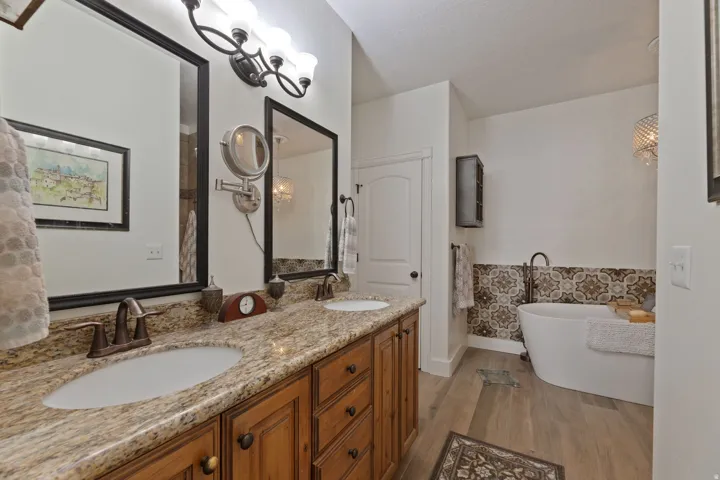Dual vanity with granite countertop and bronze fixtures, featuring a freestanding soaking tub with patterned tile surround and a wood-finish flooring