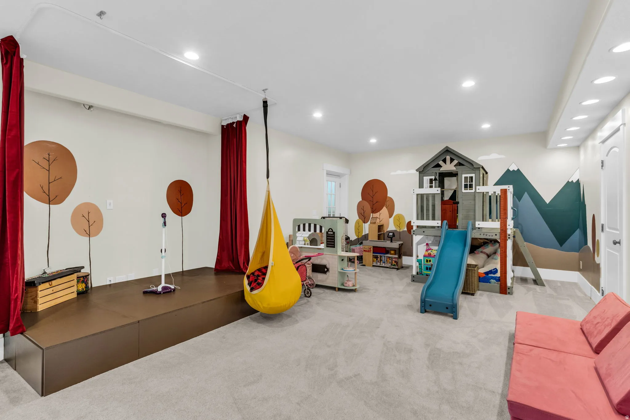 Spacious recreation room featuring light gray carpeting, recessed ceiling lighting, and a raised stage area with red curtain accents