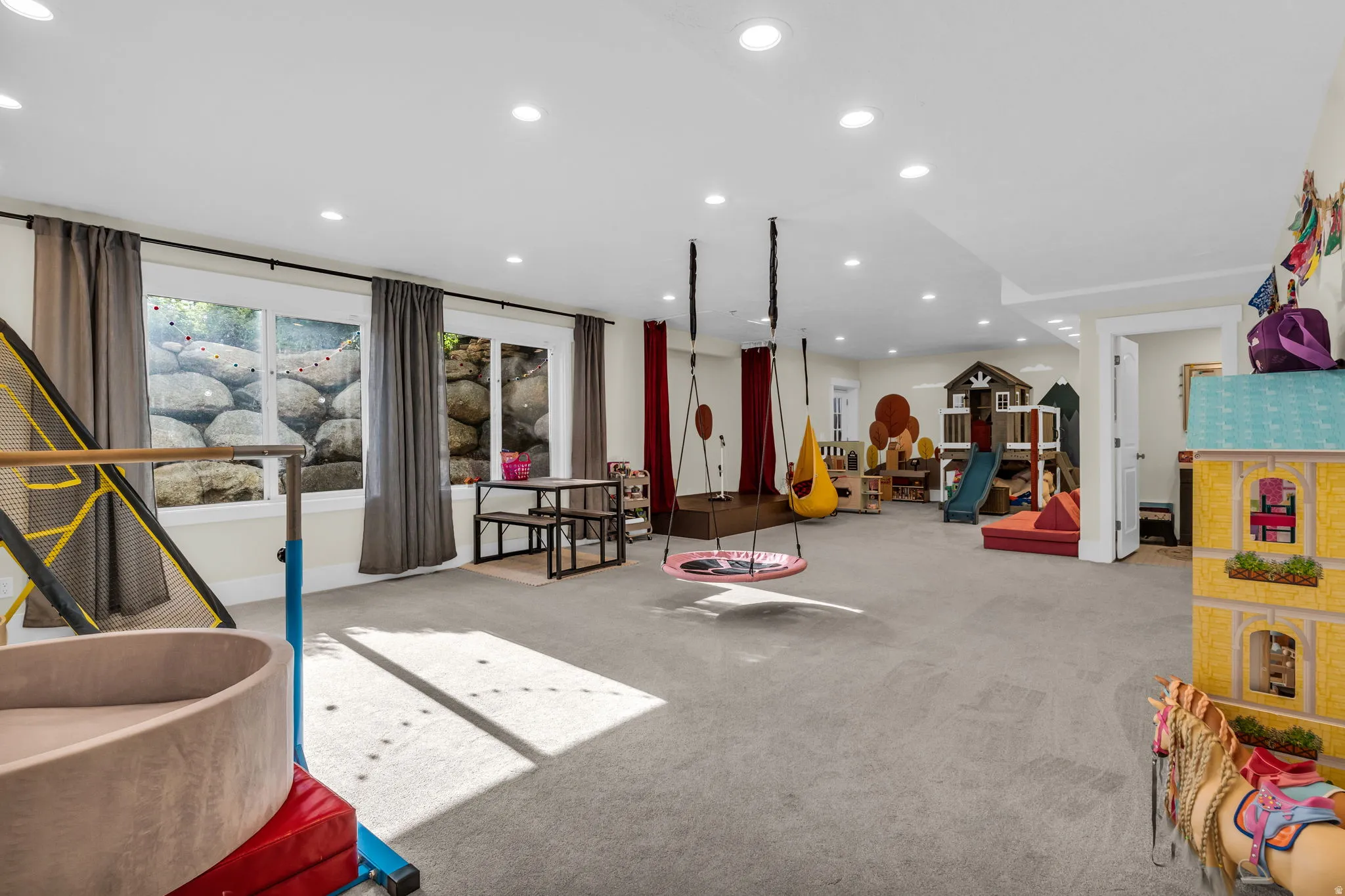 Spacious lower-level room featuring light gray carpeting, recessed lighting, and multiple windows with exterior rock landscaping
