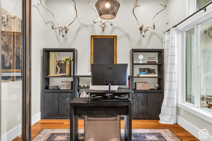 Dedicated workspace featuring wood-finish flooring, built-in shelving units with lower cabinetry, a large window with white trim, and a dark-toned suspended light fixture