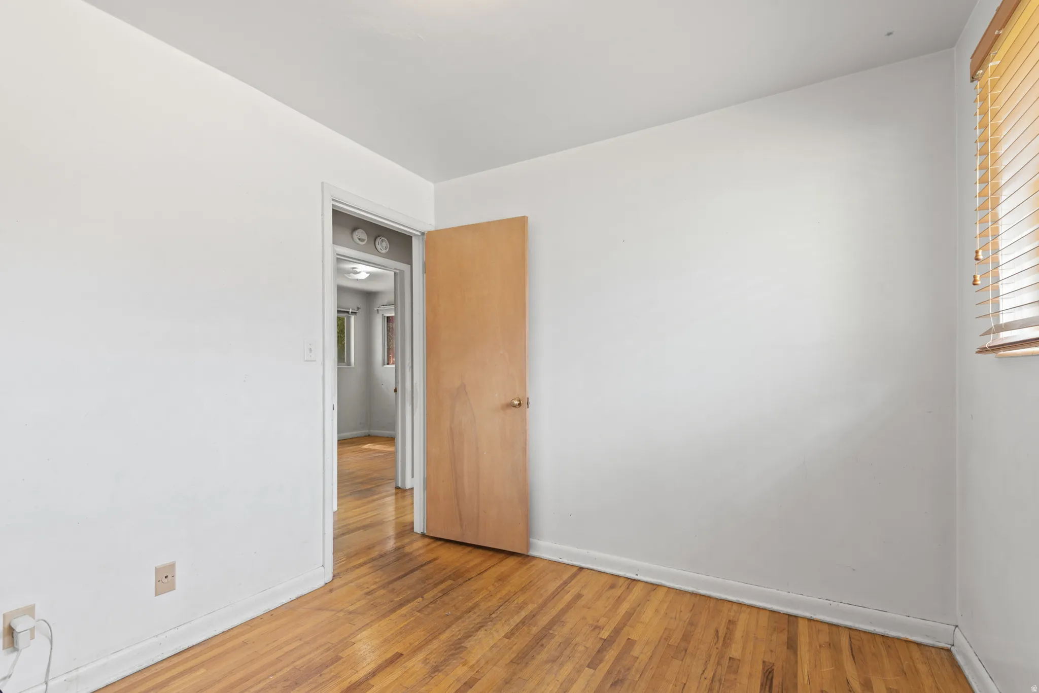 Room with wood-finish flooring, a light wood door, and a window with wood blinds