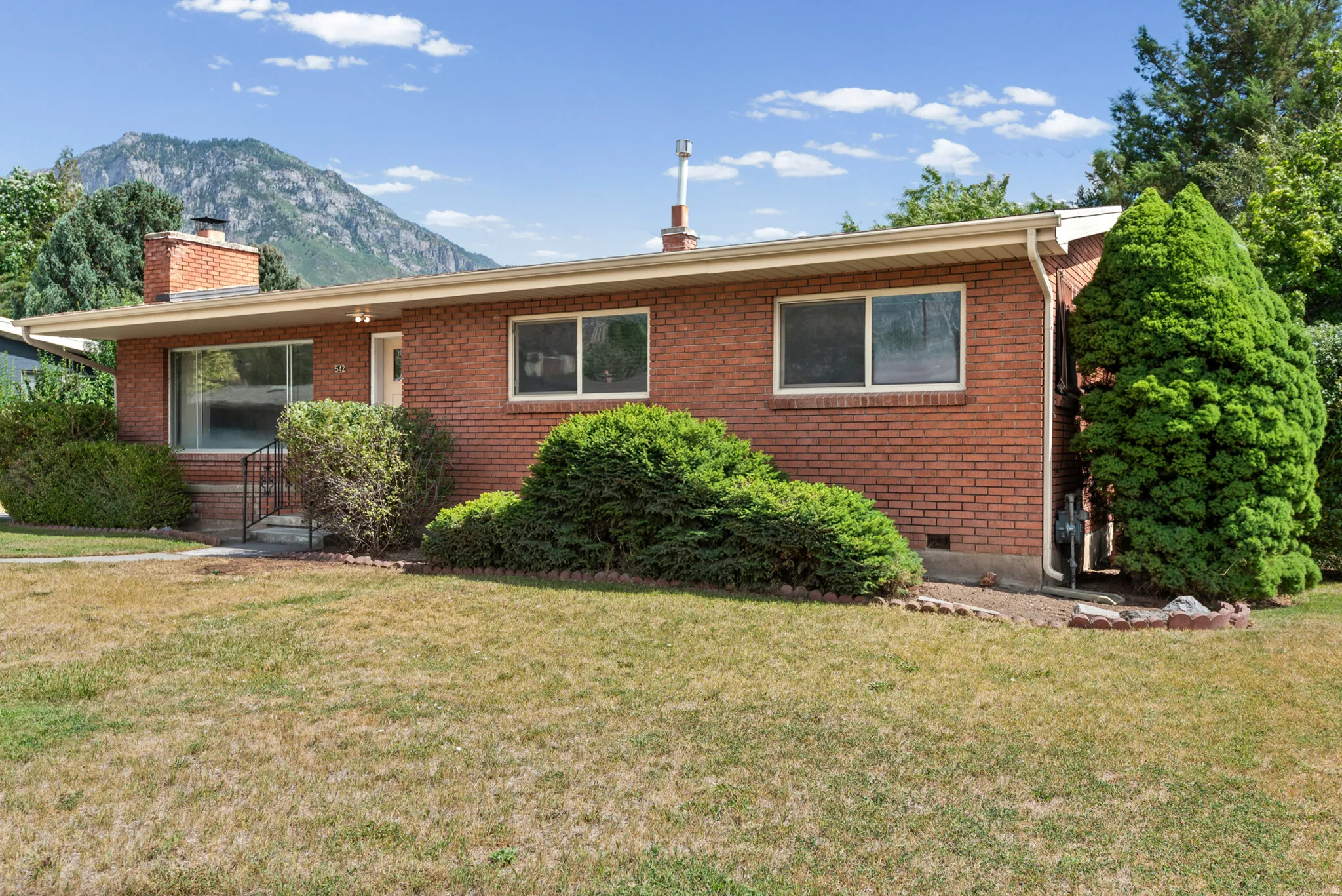 Brick exterior featuring a front porch with railing, mature landscaping, and a mountainous backdrop