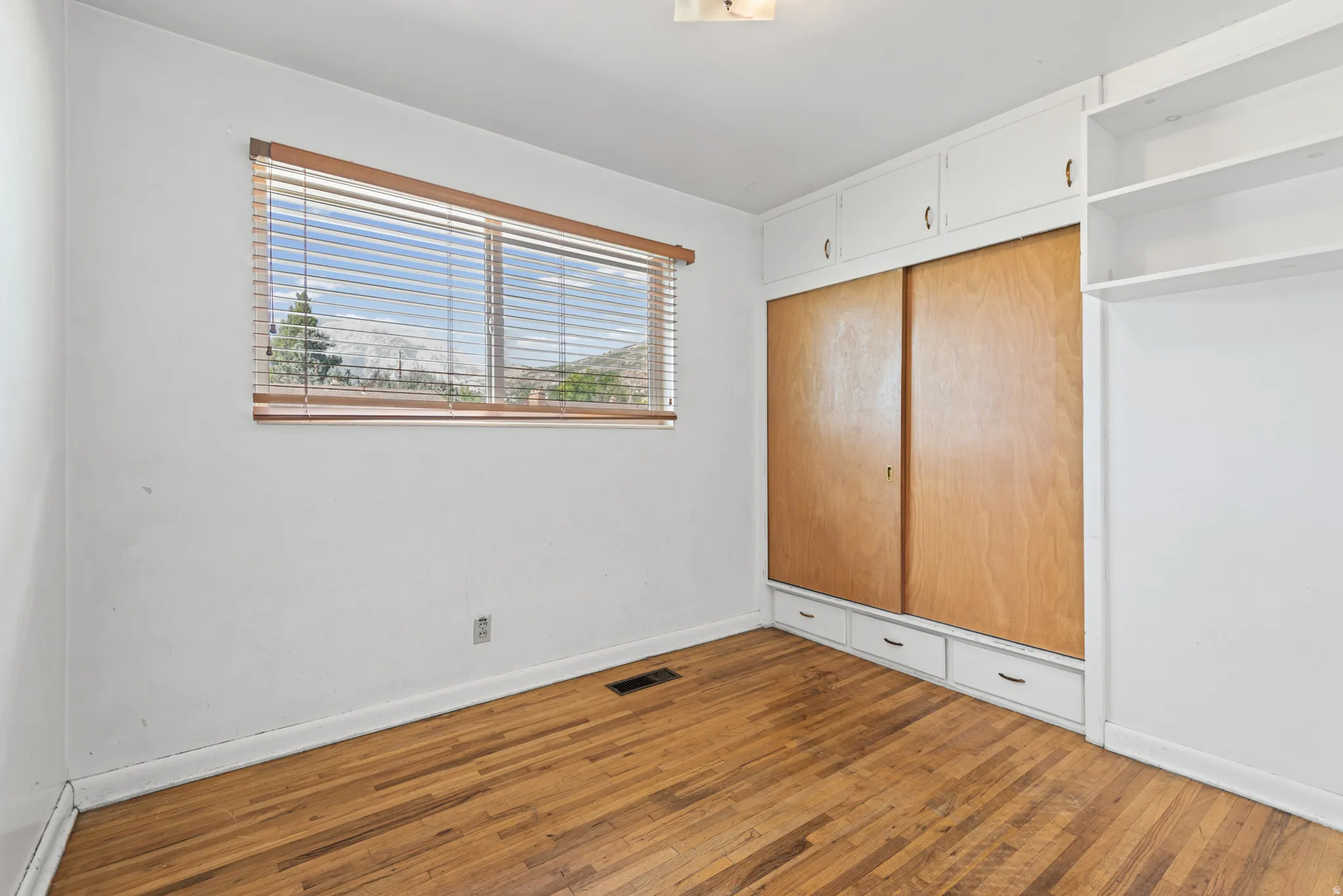 Hardwood flooring complemented by a window with horizontal blinds and built-in white cabinetry featuring wood-finish sliding doors and lower drawers