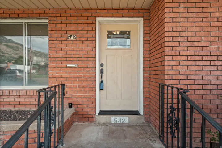 Red brick exterior featuring a cream-colored entry door with decorative glass panel, complemented by a black wrought iron railing and a window with a reflective mountain backdrop