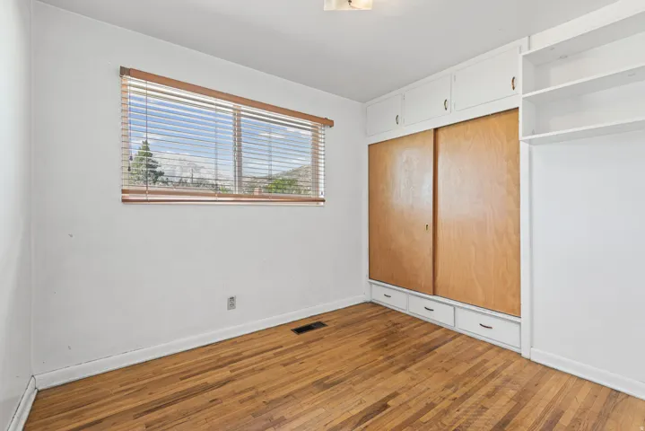 Hardwood flooring complemented by a window with horizontal blinds and built-in white cabinetry featuring wood-finish sliding doors and lower drawers