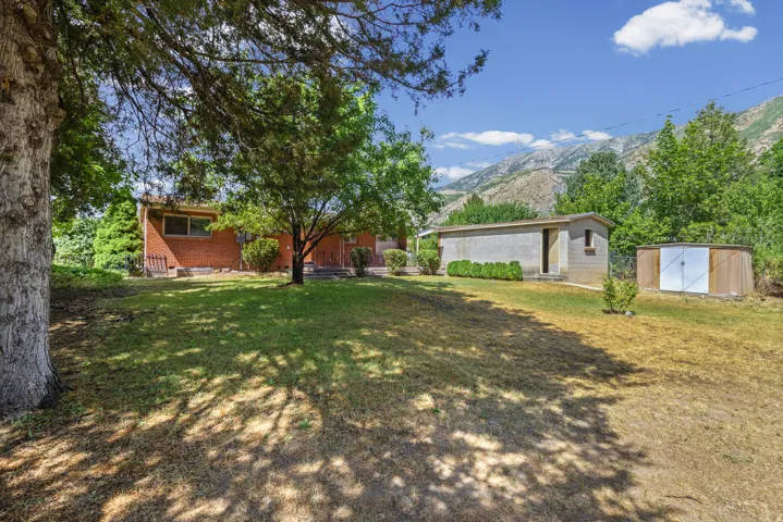 Expansive backyard featuring mature trees, a red brick exterior, a concrete block auxiliary building, a wooden shed, and mountain scenery