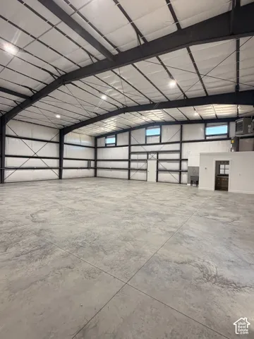 Spacious open-concept warehouse featuring a high ceiling with exposed steel beams, durable concrete flooring with a textured finish, multiple windows for natural light, and recessed lighting fixtures throughout