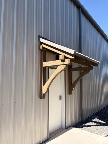 Metal-sided building exterior featuring a wood-beam awning with a light-colored shingle roof over an exterior door